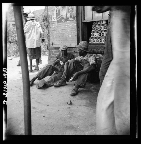 The black and white photograph captures a candid moment of three men sitting outside on the ground, possibly taking a break during work. One man is wearing a cap, another has his head down in contemplation with hands clasped together, while the third sits casually with one leg extended forward.

Behind them stands an individual in protective clothing, suggesting some form of labor or agriculture activity nearby. The setting appears to be rural, with a backdrop showing corn stalks and posters on what seems like a storage shed for agricultural supplies such as cans labeled "Hill Bros."

The presence of canned goods indicates the men might have been involved in food processing or harvesting work. A discarded cup is visible near one man's hand, hinting at casual drinking during their break.

This photograph resonates with historical context and social commentary on rural laborers' conditions. The image captures a slice-of-life moment from Georgia peach pickers, as noted by the caption provided: "Georgia peach pickers eating." This suggests that these men could be involved in agricultural labor related to picking peaches or other crops.

The photograph is credited to Dorothea Lange and sourced via Léonard Muscella. The URL hints at a broader collection of works, possibly depicting various aspects of American life during the era captured by Dorothea Lange's lens, known for her  [...]