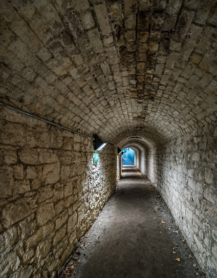 A dimly lit stone tunnel with an arched brick ceiling curves gently into the distance. Weathered limestone walls show age and water damage, with patches of discoloration. Scattered leaves cover the concrete floor, and natural light filters through openings in the left wall, revealing glimpses of blue sky beyond. A metal handrail runs along the left side of the passage.