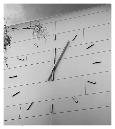 Aufnahme in schwarzweiß, Blick empor an eine Fassade einer Schule, angebracht ein riesiges analoges Ziffernblatt, bestehend aus 12 Strichen, kreisrund verteilt plus großer und kleiner Zeiger 