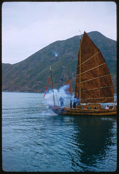 The image captures a traditional Chinese junk boat floating on the water with several individuals aboard. A large red flag is visible at the rear, and smoke billows from what appears to be an activity within or near the hull of the vessel. In the background stands a mountainous landscape under an overcast sky. The photograph bears a title that reads "Sightseeing in Hong Kong (1959)" along with a date indicating it was taken on July 1959 by photographer Toni Frissell, known for her work as a fashion and sports photographer. Additional information about the image can be found at reference number HK - X5826, Extra 1959.
