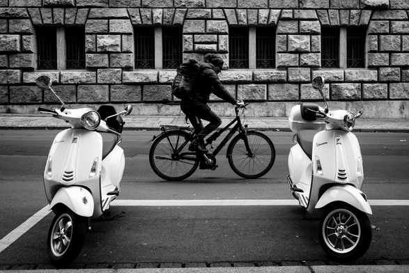The image is a black and white photograph of a street scene. The scene is dominated by a person riding a bicycle and two white scooters.

The bicycle rider is positioned in the center of the frame, moving from left to right. The rider wears dark clothing and a backpack. The bicycle is black.

On either side of the bicycle are two white scooters, parked on the street. The scooters face forward toward the viewer.
