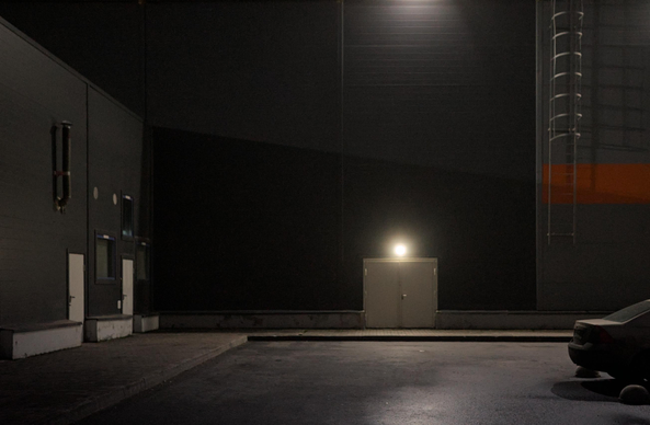 The photo shows a night scene. The wall of the hangar. A spotlight and a lamp illuminate a part of a large gray wall, in the lower part of which there is a single door. There is silence and tranquility around, which emphasizes the atmosphere of the city at night. In the foreground, you can see part of a car parked at the curb.