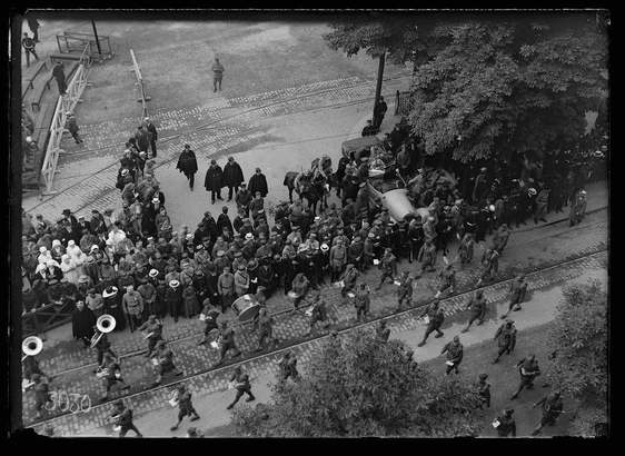 The image depicts a historical scene with individuals in military uniforms marching on a street. They are accompanied by men carrying musical instruments, such as large brass horns and drums, indicating the presence of a band. Civilians wearing hats and coats observe from both sides of the road; some wear dark cloaks or headgear suggestive of winter attire. The crowd appears engaged with the procession, suggesting an event of significance.

The setting includes trees on either side of the street, which are in full foliage, indicating it is possibly spring or summer. There's a mix of uniformed soldiers and civilians along what seems to be Avenue du President Wilson, as mentioned in the caption provided. The image captures movement with some individuals stepping over rails connected by a wire fence.

The grayscale nature suggests that this photograph could date back several decades, potentially from early 20th-century Europe or America. There's no direct indication of specific historical context within the picture itself but it is accompanied by additional information about American band and troops passing down Avenue du President Wilson along with soldiers being unloaded onto a train in another image found on the provided link.

This scene may have been part of a military parade, possibly related to an event involving wounded soldiers as mentioned in the caption. It reflec [...]