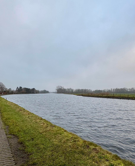 Een landschap met een kalme rivier de Vliet tussen Voorschoten en Leidschendam die door het landschap stroomt, de oevers begroeid met grasland en bomen op de achtergrond onder een bewolkte lucht.