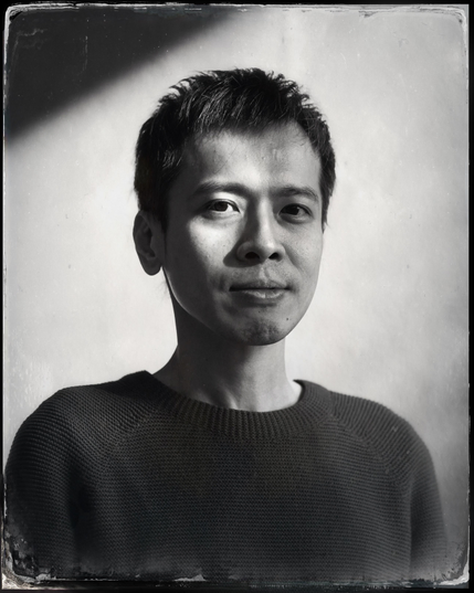 Black and white head and shoulders portrait of a young man with dark hair. He is turned slightly to the right and has the hint of a smile on his face. He is wearing a dark t-shirt. Light is falling diagonally across his face, illuminating it unevenly so that one side is more brightly lit than the other. The plain background is also lit diagonally.