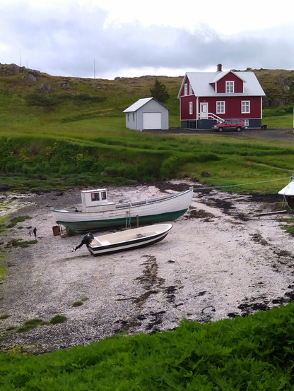 Una piccola casa di pescatori, rossa e bianca, nel nord dell'Islanda. Sulla parte bassa due barche in secca . Erba verde intorno.