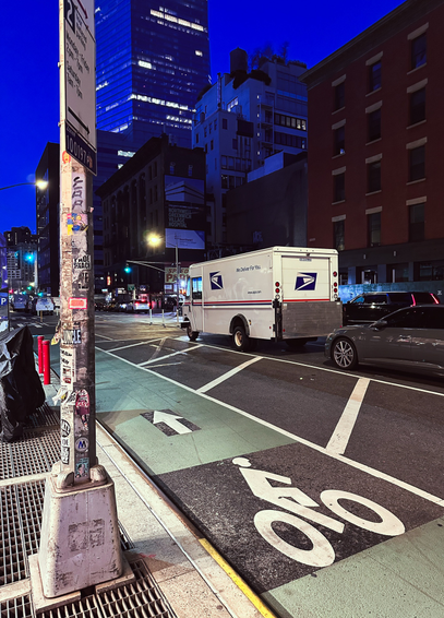 A mail truck parked next to a bike lane on West Broadway in NYC. The sky above is a deep dark blue