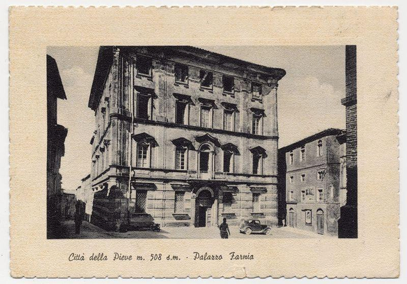 This black and white vintage photograph depicts a classical European building. The structure is characterized by its symmetrical facade, with multiple windows aligned in vertical rows on the first two floors. Architectural details such as cornices at the top of each floor add to the grandeur.

On one side, an arched doorway serves as an entryway and above it there appears to be a balcony or gallery supported by columns. The building's design suggests a significant historical context, possibly dating back several centuries given its architectural style which is reminiscent of Renaissance architecture commonly found in Italy.

The lower part of the image shows what seems like a street scene with two individuals walking on the pavement and an old-fashioned car parked at the curb. This indicates that the photo was likely taken during a time when automobiles were becoming more prevalent, possibly around mid-20th century or earlier.

There is text in Italian written across the bottom of the photograph which translates to "Città della Pieve - Palazzo Farnia" and below it there's another line indicating its location as being at via del Popolo 508 d.m. This provides a specific geographical context, identifying this building within Città della Pieve.

The overall condition of the photograph suggests age with slight wear around the edges, which is typical for vintage postcards or ph [...]