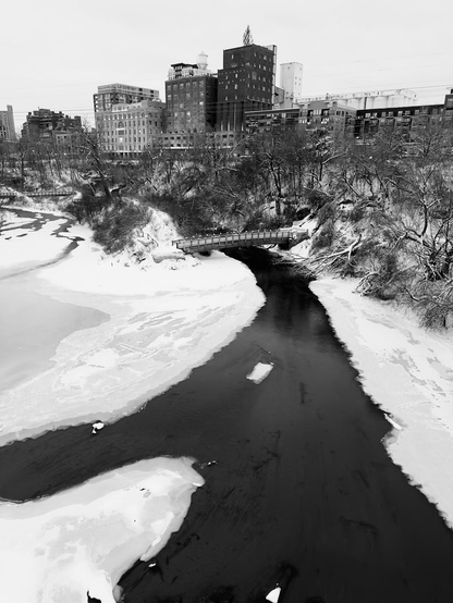 A black and white photograph shows a river flowing under a small footbridge. Open water under the bridge is surrounded by snow covered ice. Another footbridge is off in the distance and a row of buildings rises along the horizon.