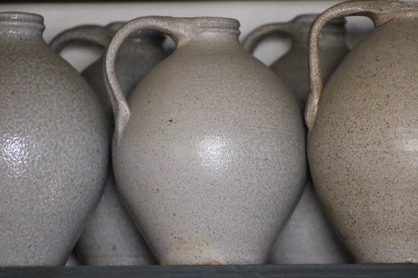 A collection of ceramic jugs with textured surfaces, displayed on a shelf. The jugs vary slightly in color, primarily in shades of gray and beige.