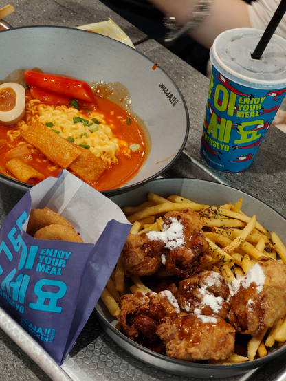 A tray of Korean comfort food, including a bowl of spicy tteokbokki with ramen, fish cake, and a soft-boiled egg, next to a plate of fried chicken and fries drizzled with sauce and powdered seasoning, plus a cup of soda.