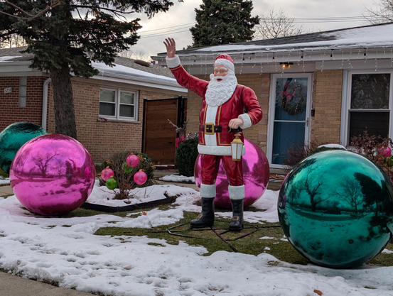 A large, Santa Claus lawn statue stands on a patchy, snow-covered front yard, surrounded by oversized shiny pink and teal ornament balls in front of a one-story brick house decorated with icicle lights.