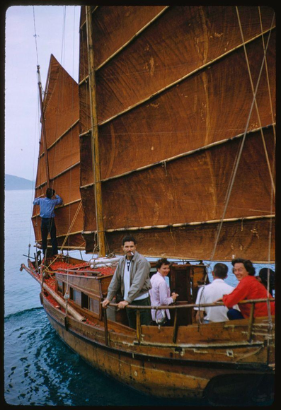 A group of people in a boat with large brown sails. The sailboat is on water and the background shows mountains or hills, indicating it could be near shore. Onboard are five individuals: one man standing at front holding part of the ship's rigging while smoking a cigarette; two women seated side by side smiling at camera wearing pink and red blouses respectively; another woman in white blouse to right with hand on railing looking out towards water; finally, back row is partial view of person sitting. This photograph shows an early travel or tourism moment for Hong Kong as part of its colonial era under British Crown Colony from 1957-1997 located southern coast China