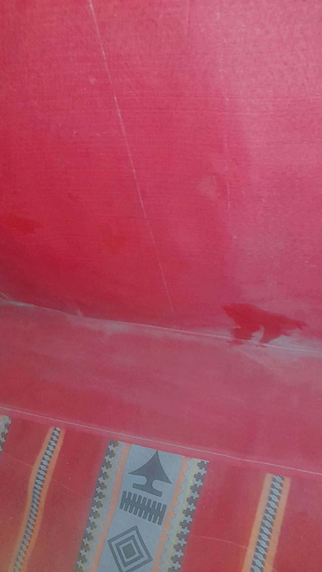 Red tent interior with a patterned design at the bottom edge. The upper portion has a smooth, textured appearance with a few imperfections, and the lower edge features a rectangular design with repetitive geometric patterns. We can see it's wet because of the rain.