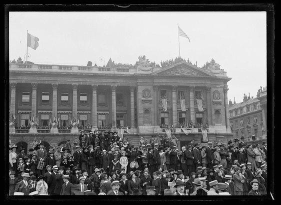 The image depicts a historical scene, likely from around the early 20th century. It is black and white, which suggests it's an old photograph or drawn to resemble one. The setting appears to be Place de la Concorde in Paris during celebrations for American Independence Day (4th of July). A large crowd has gathered on the street, with individuals dressed in period clothing such as suits, hats, and military uniforms indicating a formal occasion.

In the background is an opulent neoclassical building adorned with columns and statues. The structure's design suggests it could be a government or historical landmark. Two flags are prominently displayed atop: one appears to be the French tricolor (blue, white, red), while the other seems to represent America based on its stars and stripes.

The people in the foreground seem engaged, perhaps watching something of interest out of frame or possibly waiting for an event such as a parade. Some individuals hold signs with text that is not legible from this distance, which might have been used during political demonstrations common at public celebrations around that time period. The overall mood seems festive and communal.

Notable features include the density of people indicating significant attendance for the occasion; the diverse attire suggesting various social classes or roles in society were represented among attendees; and the cl [...]