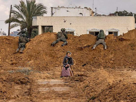 A photograph of a woman sitting on a chair behind a bank of dirt, where three zionist genocidaires are lying.
