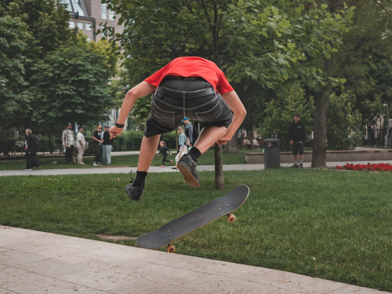 Ein junger Mann mit Skateboard in einem Park. Er hängt hoch in der Luft, unter ihm das Skateboard - auch in der Flugphase.
Der junge Mann ist von hinten zu sehen, Beine angezogen.
Im Hintergrund einige Menschen beim Spazieren gehen.
