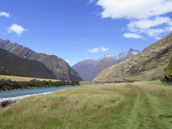 A picture taken on the South Island of New Zealand.  The picture shows mountains in the distances flanked by steep hilly areas on either side (the hilly area on the left is densely covered with trees while the hilly area on the right is mostly barren).  There is a large grassy meadow in the foreground with a stream running through it.