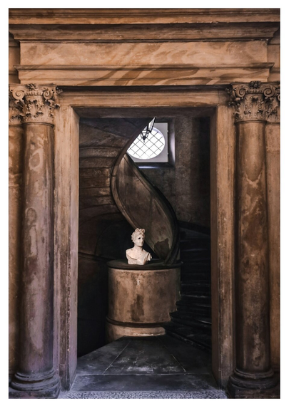 This photo is of classical architectural scene with a focus on a doorway leading to a spiral staircase. The doorway is framed by two ornate columns, and through the doorway you see a marble bust standing at the base of a curving staircase. There is soft natural light which filters through a round window at the top of the staircase.