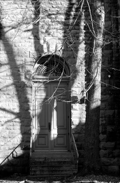 A black and white photograph of a double wooden door surmounted by an arched window, at the top of a six-step concrete staircase with metal railings, set in a rectangular stone wall. A wall sconce is mounted on the wall to the right of the door. A large tree with a thick, straight trunk grows very close to the wall to the right of the door. Its bare lower branches are also visible. The sun casts the shadow of the tree and its branches onto the door and the wall.

Photographie noir et blanc d'une porte double en bois surmontée d'une fenêtre en arche, en haut d'un escalier de six marches en béton avec des rampes en métal, dans un mur de pierres rectangulaires. Une applique murale se trouve fixée au mur à droite de la porte. Un grand arbre au tronc épais et droit pousse très près du mur à droite de la porte. Ses branches inférieures dénudées sont aussi visibles. Le soleil projette l'ombre de l'arbre et des branches sur la porte et le mur.