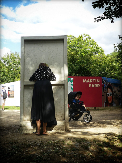 Une femme passe la tête dans un trou pour faire une photo. Son fils dans une poussette la regarde. C'est dans le cadre d’une exposition de photos de Martin Parr