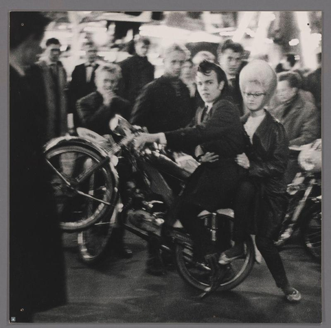The image is a black and white photograph capturing an urban scene with multiple people. In the foreground, there's a woman on a motorcycle wearing what appears to be a fur coat or wrap around her shoulders. She has distinctive hair styled in large volume, possibly blond, which adds a striking contrast against the dark tones of her clothing. The focus is not clear enough to discern detailed expressions; however, she seems poised and attentive.

Behind her are several individuals who seem caught off-guard by something out of frame or camera's perspective. One man directly behind her looks in the same direction with an alert expression while another nearby has a more neutral gaze. Further back, there appears to be a gathering of people, possibly spectators or bystanders, contributing to a sense of bustling activity.

The composition and attire suggest this could be from mid-20th-century urban life, though no specific identity markers are visible that would pinpoint the exact location or event taking place at the time. The image lacks any explicit context but seems to capture candid moments in public space with an artistic flair given its monochrome aesthetic and apparent intentionality behind composition.

It is important not to speculate on identities or make assumptions about individuals beyond what can be visually confirmed, adhering strictly to observable facts within t [...]