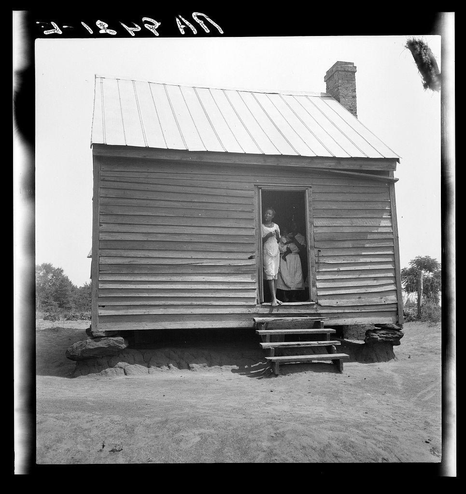 The image depicts an old, rustic wooden cabin with a sloped metal roof and exposed beams. A young girl stands in the doorway of this structure, her hands clutching onto the door frame as she peers outwards. She appears to be wearing a light-colored dress or skirt. To the right side of the cabin is another person standing at an open window ledge within the main living space; only their lower body and legs are visible from inside.

The building seems humble with its construction, possibly indicating modest living conditions in rural Georgia as described by Dorothea Lange's photograph. The ground surrounding the structure appears to be a dirt path or clearing, suggestive of farm-like surroundings where pickers might reside during harvest season.

A handwritten caption on top reads "J-1257P AD," which may refer to a reference number for this specific photo within an archive collection. Additional information about this image can indeed be found in the provided source link: https://images.loener.nl/DorotheaLange/full/6495/6495b84aba25398d90c50432.jpg