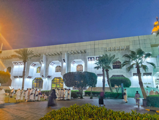 ​A wide-angle landscape photograph taken at twilight shows a large crowd of people, mostly men wearing white ihram clothing and some women in black abayas, standing on a wet paved walkway outside a massive white stone building. The building features modern geometric window designs that are brightly illuminated from within. Several palm trees and manicured green hedges line the path. The sky above is a deep gradient of twilight blue and purple, with a warm glow coming from the building lights.