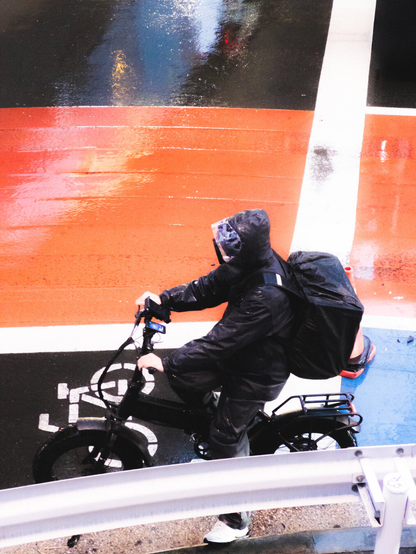 A person wearing a black raincoat rides a bicycle on a wet road marked with bike lane symbols. The road is glossy from rain, reflecting light, with sections painted red and white. The cyclist carries a large backpack. A metallic guardrail is visible at the bottom of the image.