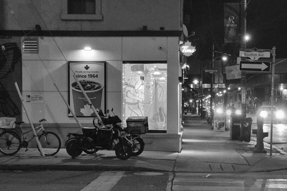 Night. At left is the side of a Tim Hortons, its one window brightly lit in the dark. Parked outside are two unattended food courier bikes, waiting. At right is Queen Street itself, full of the lights of cars, the train overpass above.