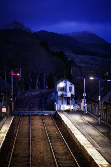 Der Bahnhof Blair Atholl in Schottland ist bei Einbruch der Dunkelheit abgebildet. Beleuchtete Bahnsteige und das charakteristische Stellwerk bilden einen Kontrast zum tiefblauen Himmel und den dunklen Bergen im Hintergrund. Die Gleise verlaufen dynamisch in die Tiefe und strukturieren die Komposition.