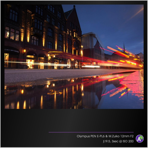 a color outdoor photograph. a bus passes in front of a row of 17th-century buildings and warehouses on a cobblestone street. there is a puddle in front of the bus that reflects the scene. it is almost dark, so the lights of the buildings and the bus figure prominently in the photograph. there are no people or animals in this picture.