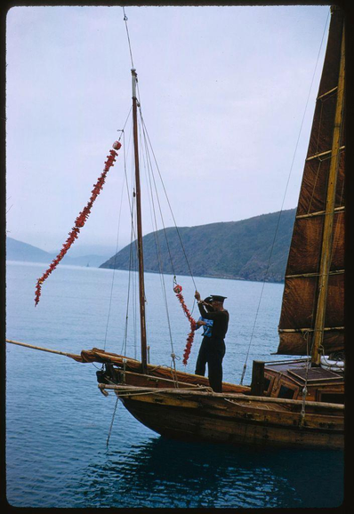 A sailboat on water near mountains is decorated in red, with a person standing at the front of the boat. The person appears to be adjusting or hanging decorations from one side of the mast and has their right arm extended out toward it. There are various ropes and rigging seen around them as well as attached to parts of the sailboat structure including what looks like a bamboo pole extending off into the water on the left hand side, which is tied down with rope at its end.