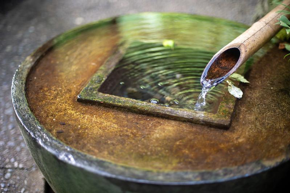 Water trickles from a bamboo pipe into a shallow circular basin with a deeper square central section in the middle.