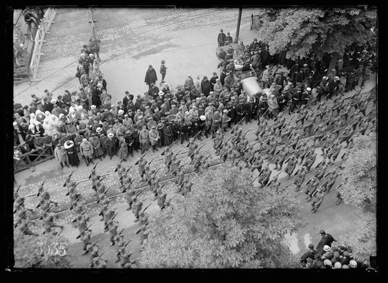 The image depicts a large group of soldiers marching in formation during what appears to be an official event or parade. The scene is set on Avenue du President Wilson, as mentioned in the provided context about Paris's Fourth of July celebration. In this black and white photograph taken by Lewis Hine, American infantry members are prominently featured marching down the avenue.

Uniformed men hold rifles at attention while a mix of civilians including women wearing hats observe from both sides of the parade route. Some individuals appear to be wounded soldiers seated in an automobile on the right side of the frame, highlighting their presence and participation despite injury. The atmosphere is formal yet somber due to the wartime context.

The photo's timestamp suggests it was taken around July 1918 during World War I when many European countries hosted parades for American troops as part of a show of support or celebration amidst ongoing conflict. This photograph captures both military discipline and civilian interest, providing insight into how soldiers were viewed by onlookers in the context of wartime camaraderie.

Please note that this description is based solely on visual elements present within the image and does not include any speculative interpretations beyond what can be directly observed.