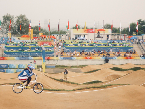 A color photo of a BMX rider in mid-air on a dirt track at the 2008 Olympics. The track is made of dirt hills with a white striped dividing line. In the background, spectators watch from tiered seating, some under umbrellas, while more people stand near the track. The front of the stands has a long banner in shades of red, yellow and blue.  From the back of the stand are many flags from various countries. The atmosphere of the photo is quite light because it was a summer day in Beijing and very hot and humid