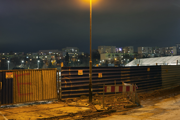 Nocna fotografia. Na pierwszym poście widać metalowy płot, a przed nim stoi latarnia oświetlająca wjazd na budowę. W zasadzie widoczny jest jedynie słup starej latarni. Wjazd na budowę wyłożony jest betonowymi płytami, a przy nim, po prawej stronie jest niewielki, ogrodzony biało-czerwoną barierką kawałek ziemi. Nie widać co jest za ogrodzeniem, sponad niego wyłania się osiedle mieszkaniowe z wielkiej płyty - okna w blokach świecą, a po prawej stronie, tuż ponad płotem widać jakiś jasny, jakby oświetlony od środka budynek. Nad wszystkim jest niebo - nocne, ale nie całkiem ciemne, w środku, u samej góry kadru, na słupie rozjaśnia je pomarańczowe światło latarni.