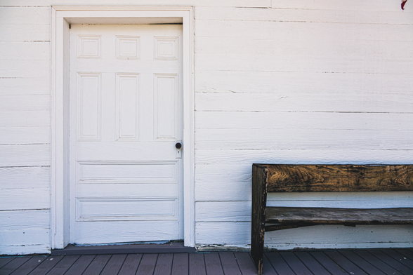 A white-painted wooden building exterior featuring horizontal lap siding with weathered, peeling paint showing age and wear. On the left is a white door with an eight-panel design (six rectangular panels arranged in two rows of three, plus two horizontal panels at the bottom) and a black round doorknob. To the right of the door sits a rustic wooden bench with a natural wood finish showing pronounced grain patterns and darker weathering. The bench has horizontal slats for the backrest and seat. The structure has a dark-painted wooden porch floor. The overall composition shows signs of aging and rural character with cracked paint and worn surfaces throughout.