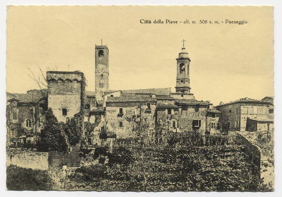 The black and white photograph depicts a quaint European town setting. Two prominent bell towers stand in the center, one featuring an arch-shaped window below its belfry and another with a cross atop it, indicating religious significance possibly as part of a church or cathedral structure. Surrounding these central structures are various buildings that appear to be residential houses, characterized by their rustic stone facades, pitched roofs, and small windows typical of historical European architecture.

The town is nestled amidst vegetation; vines seem to cascade down the sides of some walls, while trees with sparse foliage suggest a temperate climate or possibly a season where leaves have fallen. The overall scene gives an impression of tranquility and timelessness, capturing what may be Città della Pieve in Italy as mentioned on the photograph's caption "Città della Pieve - alt. m. 508 s.m., - Paesaggio".