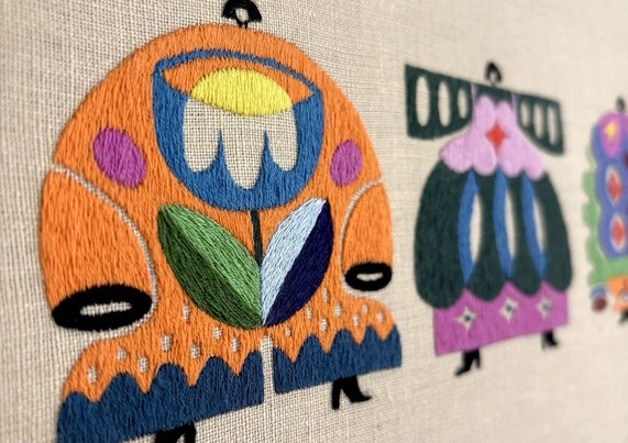 Colorful embroidered figures with floral and geometric designs on a light fabric background, showcasing artistic textile craftsmanship.