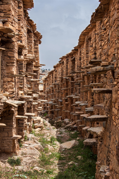 Narrow pathway through ancient stone ruins with multiple tiers of exposed rooms and ledges, no roof present, under a partly cloudy sky.