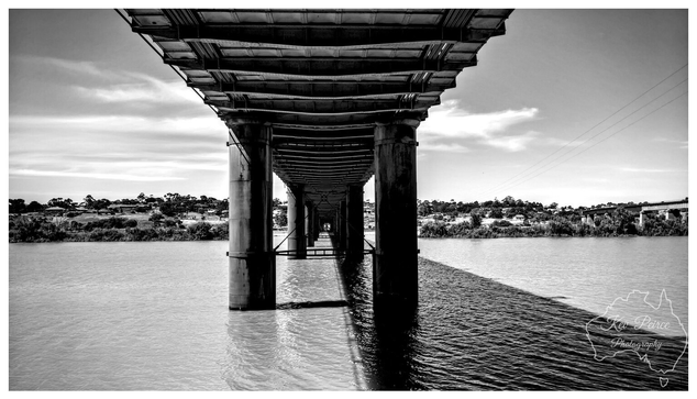 Black and white low angle photograph taken from under the historic Murray Bridge railway bridge in South Australia, showing a long, receding perspective of the massive concrete support piers standing in the Murray River.  The dark, textured underside of the bridge structure looms overhead, creating a strong contrast with the bright sky and the reflected light path on the water. Signed Kev Peirce Photography.