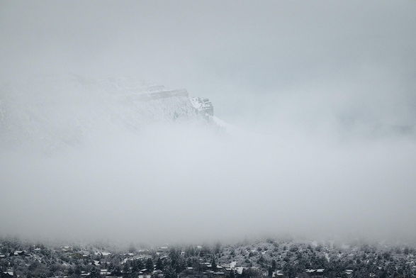 Sparsely-forested and snow-blanketed mountain slopes and cliffs peek out of dense winter clouds sitting low in a valley with snow-covered homes and trees revealed only in the foreground. 