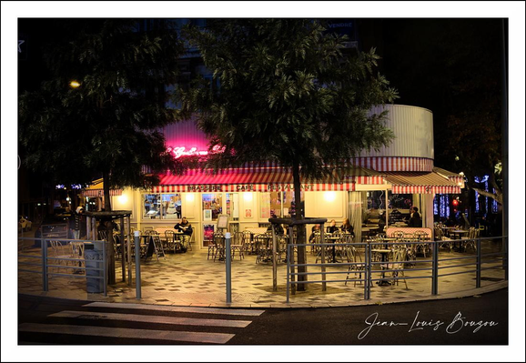 A small corner café sits quietly under the night sky, its rounded facade bathed in a warm honey glow that spills onto the sidewalk. A striped fabric awning wraps the building, and a row of pendant lights and soft interior lamps give the windows a welcoming, intimate light. Rows of metal bistro tables and chairs fill the tiled patio — neat, empty, and arranged as if waiting for the next wave of customers — while low bollards and a zebra crossing in the foreground anchor the scene to the street. Above the entrance a neon sign throws a rose-pink halo into the leaves of nearby trees, adding a splash of color against the deeper blues and blacks of the night. Trees and hanging branches frame the café, their shadows and dappled light softening the hard geometry of the building and pavement. The checkerboard floor pattern under the tables gives the space a retro, timeless feel, and small details like the menu board near the door and a curtained window hint at the personality of the place. Overall the image feels quiet and slightly cinematic — a calm, inviting pocket of light and warmth in an otherwise dark city night. 
