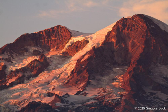 Red-saturated sunset light angles across the summit of a ragged glacier carved peak, painting jumbled ridges of snow, ice, and shattered rock.