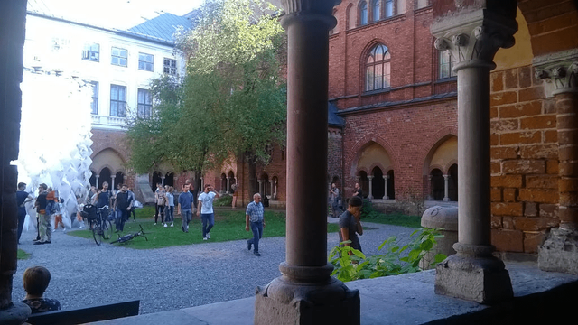 Photo of a center square inside of a churches territory, looking through the glass-less windows, with a plastic object off to the left of the photo, with people milling around the open square.