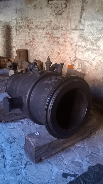 Photo of an old bell placed on wooden planks to keep off the ground, near a yellow and red brick wall, with decorative metal and wood elements around and on the ground.