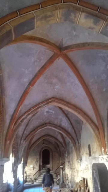 Photo of an old church hallway, yellow and red bricks and decorative coloring on the ceiling, light shining through the windows, with small stairs at the end of the hallway with a seemingly-metal door. Metallic and wooden decorations are by the wall.