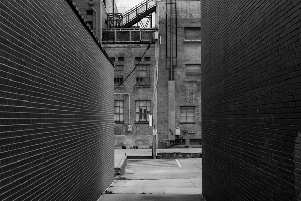 A black and white photo of an alleyway between tow red brick buildings which leads to a much larger back alley downtown.