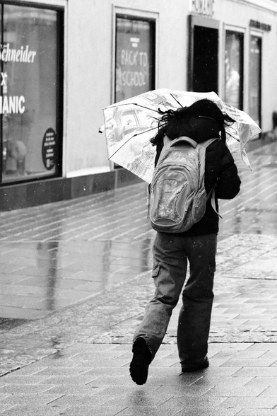 Das Foto zeigt eine Straßenszene in Schwarz-Weiß in der St. Pöltenter Innenstadt an einen regnerischen windigen Tag. Man sieht eine Person mit Regenschirm der durch den Wind verbogen ist.